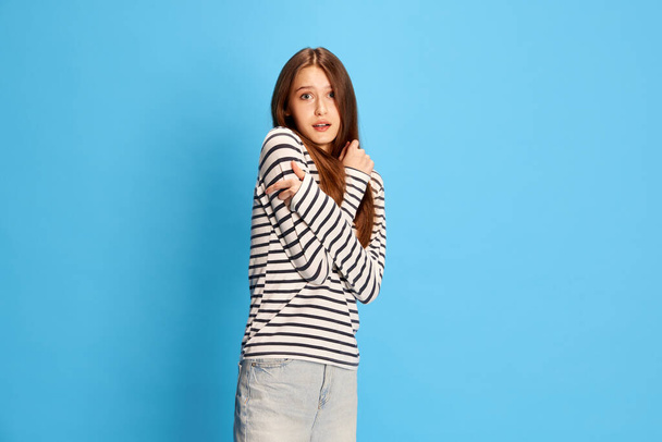 Portrait of young attractive girl in casual clothes, hugging herself, feeling fear and stress against blue studio background. Concept of human emotions, fashion, youth, lifestyle, female beauty, ad - Foto, Bild