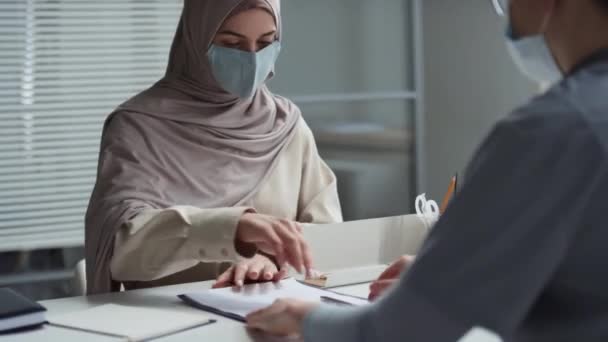 Молода мусульманська пацієнтка в хіджабі та захисна маска підписує медичний документ у буфері після консультації та тремтячої руки лікаря, сидячи за столом - Кадри, відео
