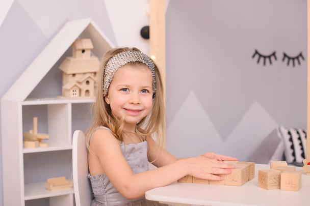 ein Mädchen mit einem Reifen im Haar spielt Würfel im Kinderzimmer, sitzend und stehend an einem weißen Tisch - Foto, Bild