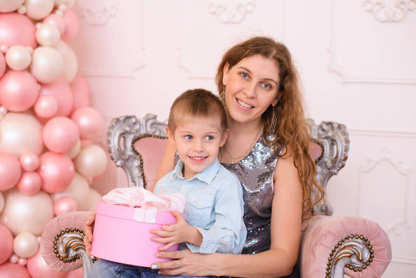 Мама з довгим хвилястим волоссям, тримає сина на руках, а рожева подарункова коробка з білою стрічкою - Фото, зображення