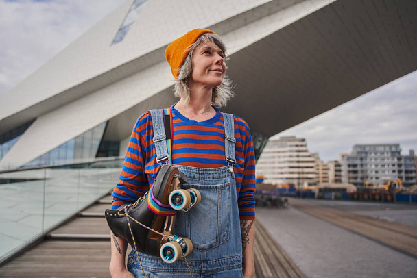 Positive adult woman wearing bright clothes holding roller skates and looking away while standing over urban architecture. Sport and hobby concept - Fotografie, Obrázek