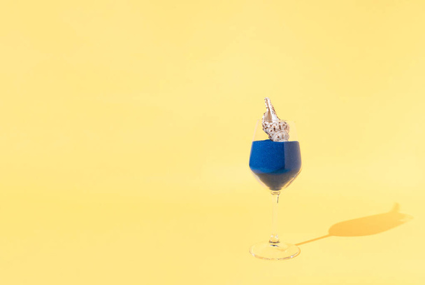Abstrakti comopositon valmistettu lasista täynnä sinistä hiekkaa simpukka kirkkaan keltaisella pohjalla. Summer beach reresresreshment, tai trooppinen loma käsite kopioi tilaa. - Valokuva, kuva