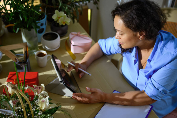 Γενική άποψη των γυναικών ανθοπώλης, floral designer sketching on digital tablet, εργάζονται σε ανθοσυνθέσεις σε ένα στούντιο ανθοκομίας, κάθεται στο τραπέζι με πρόχειρο, που περιβάλλεται από λουλούδια και φυτά - Φωτογραφία, εικόνα