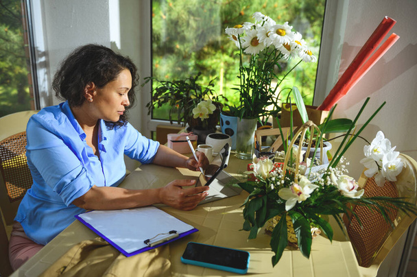 Afro-Amerikaanse mooie zwangere vrouw trekt op digitale tablet, schetsen, zitten aan het bureau, omgeven door verse bloemen en planten in een bloemen design studio. Arrangementen en geschenken. Kleine bedrijven - Foto, afbeelding