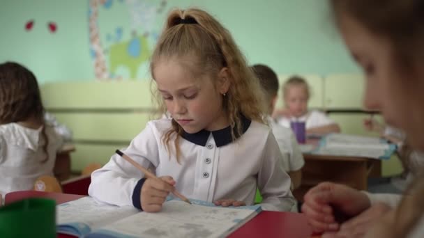 Cute smart elementary school girl learns to write, do homework, sitting at the table, charming pretty preschool child learns by taking notes, childrens elementary education concept - Footage, Video