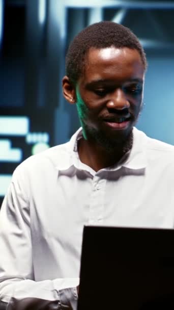 Verticale video Gelukkig Afrikaans Amerikaans werknemer met behulp van laptop om het energieverbruik te beoordelen over serverkasten componenten. Werknemer zorgt ervoor dat datacenter elektronica temperatuursensoren werken - Video