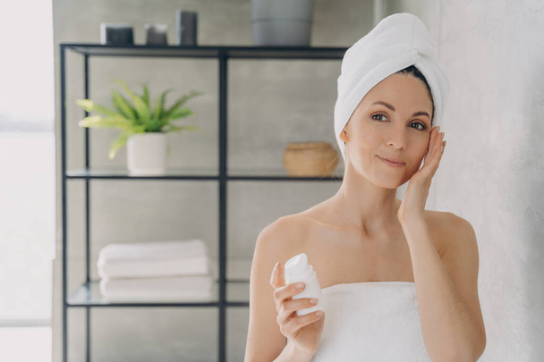 European lady applies face lotion, attractive girl with towel after bathing. Spa resort, face moisturizer, lifting cream. - Photo, Image
