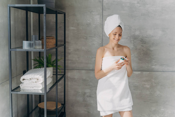Ευρωπαϊκή κυρία εφαρμόζει κρέμα από βάζο. Ελκυστικό κορίτσι με πετσέτα μετά το μπάνιο. Ευτυχισμένη γυναίκα κάνει ντους στο σπίτι. Έννοια φροντίδας δέρματος. - Φωτογραφία, εικόνα