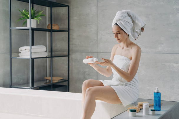 A beleza europeia envolve na toalha, aplica a nata de corpo depois do banho. Chuveiros de mulher, manhãs em casa. Anti-celulite, massagem, cuidado do corpo. - Foto, Imagem