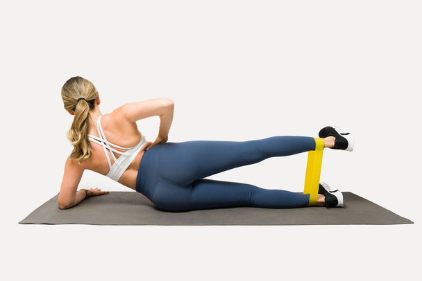 Vista posteriore di una ragazza in forma atletica con leggings che lavora con bande di resistenza e allenamento sul tappeto da ginnastica - Foto, immagini