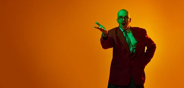 Portrait of young bald man with moustache in suit, emotionally talking against orange studio background in neon light. Astonished. Concept of human emotions, facial expression, lifestyle - Foto, Imagem
