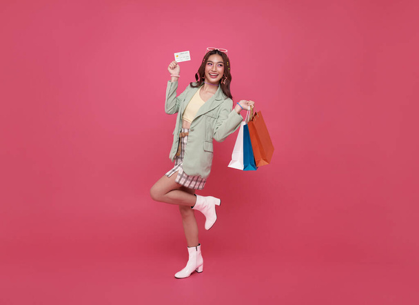 happy smiling thai asian woman enjoying shopping, she is carrying shopping bags and showing credit card isolated on pink background. - Photo, Image