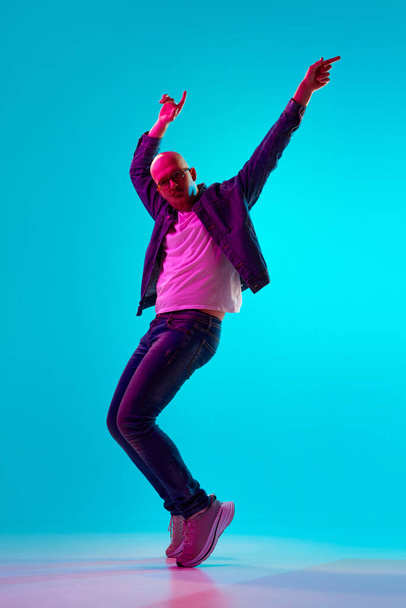 Full-length portrait of young bald man with moustache in casual clothes dancing against blue studio background in neon light. Concept of human emotions, facial expression, lifestyle - Photo, image