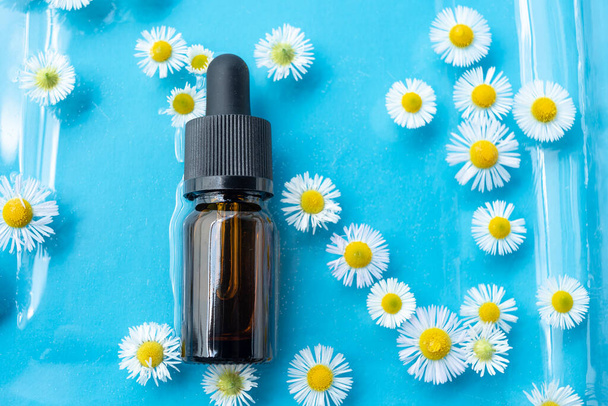 A bottle or bottle for essential oil with a pipette, lies in the water surrounded by flowers on a blue background. Background for advertising, beauty industry, cosmetology, medicine - Fotó, kép