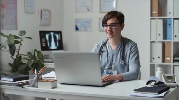 Fiatal magabiztos orvos szemüvegben és egyenruhában, aki tablettákat szed és megmutatja a betegnek az online konzultáció során, miközben a laptop előtt ül az orvosi rendelőben. - Felvétel, videó