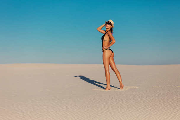 Portret van sexy mooi gelooid model vrouw poseren in de mode zwarte Badmode Bikini, Fedora hoed en zonnebril zand woestijn strand. Exotische land reizen en rest-concept. Slank figuur en sport lichaam. - Foto, afbeelding
