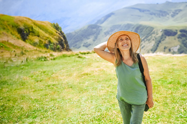 Portrait of a smiling happy elderly woman tourist traveling with a camera posing against the backdrop of mountains. Old elegant lady in straw hat on grass at countryside. Active retired people concept - Fotografie, Obrázek