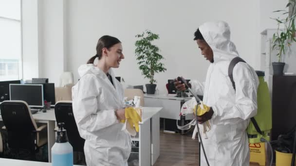 Sivunäkymä hidas liike nuori musta mies ja valkoihoinen nainen yllään suojapuvut seisoo toimistossa chattailuun päätyttyä desinfiointityön - Materiaali, video