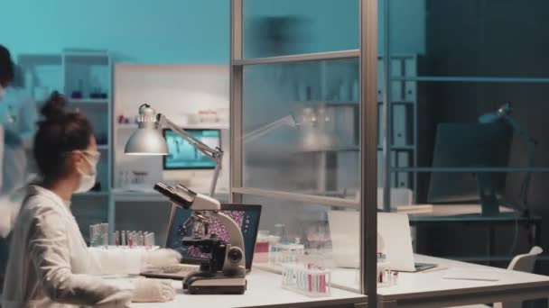 Tijdspanne van jonge vrouwelijke chemicus in laboratoriumjas, beschermend masker, handschoenen en brillen die klinisch onderzoek verrichten op de werkplek met microscoop, laptop en reageerbuizen die vloeistoffen bevatten - Video
