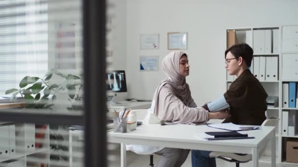 Jonge moslim vrouwelijke arts in hijab en uniform meten bloeddruk van zieke patiënt zitten voor haar door bureau in medisch kantoor tijdens overleg - Video