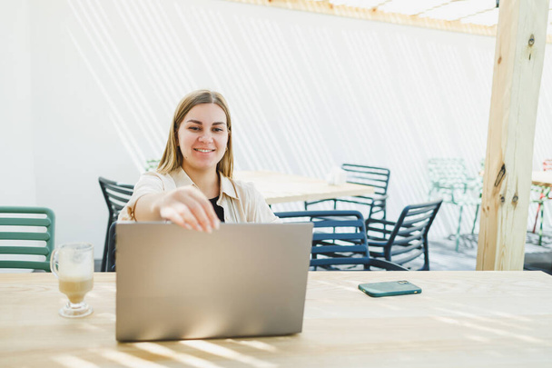 Young happy woman sitting at outdoor cafe table and talking on phone with cup of coffee, smiling woman enjoying telecommuting in cafe or studying online on laptop - Photo, Image