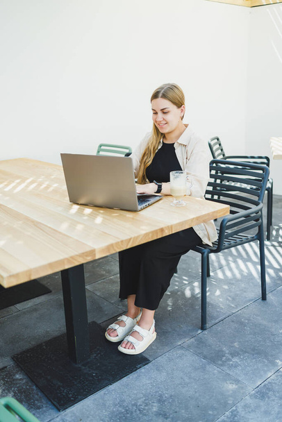 Jeune femme heureuse assise à une table de café en plein air et parlant au téléphone avec une tasse de café, femme souriante appréciant le télétravail dans un café ou étudier en ligne sur un ordinateur portable - Photo, image