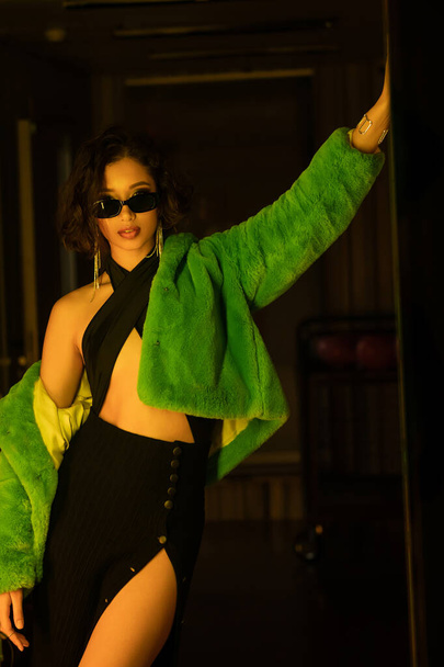 Fashionable young asian woman in dress, sunglasses and faux fur jacket posing in dark night club - Fotó, kép