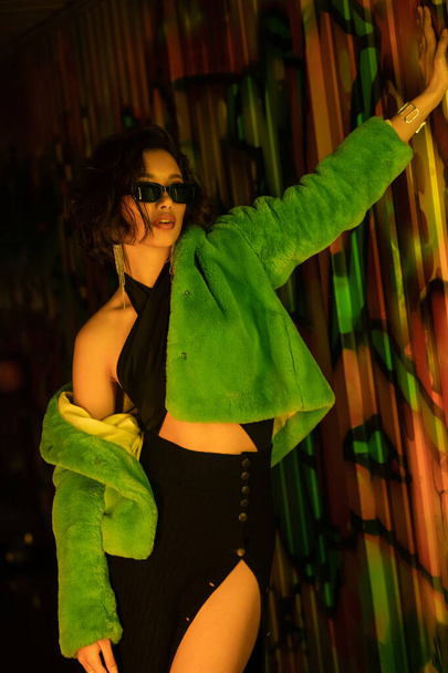 Fashionable woman in sunglasses, dress and faux fur jacket standing near graffiti in night club - Fotoğraf, Görsel