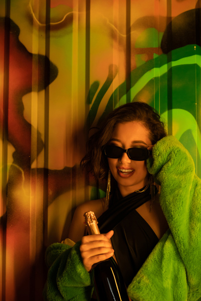Joyful and stylish asian woman wearing sunglasses and holding champagne near graffiti in night club - Photo, Image