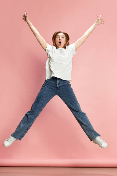 Full-length portret van jonge vrouw in casual kleding vrolijk springen tegen roze studio achtergrond. Een uitbarsting van emoties. Concept van menselijke emoties, levensstijl, jeugd, mode, reclame - Foto, afbeelding