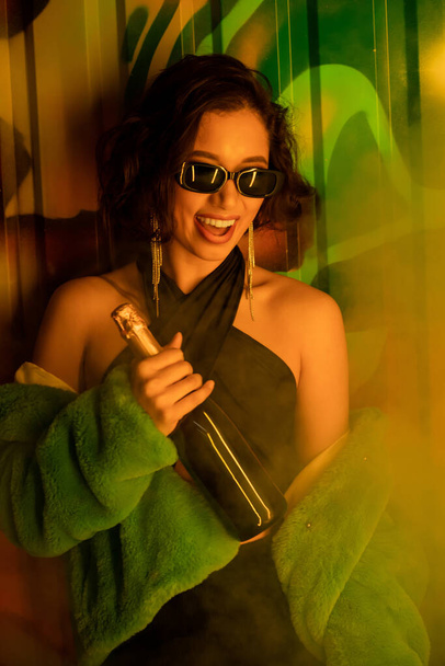 Brunette woman in sunglasses and fake fur jacket holding champagne near graffiti in night club - Foto, afbeelding