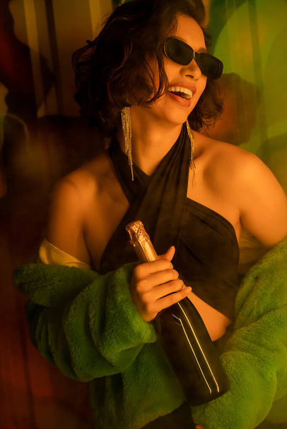 Joyful woman in sunglasses and faux fur jacket holding champagne bottle near graffiti in night club - Fotografie, Obrázek