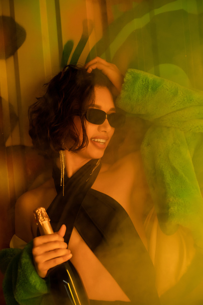ナイトクラブでグラフィティと煙の近くにシャンパンを持っているサングラスのスタイリッシュな陽気な女性 - 写真・画像