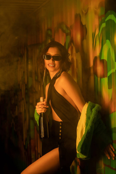 ナイトクラブのグラフィティ近くでシャンパンを持っているサングラスとドレスでファッショナブルな若い女性 - 写真・画像