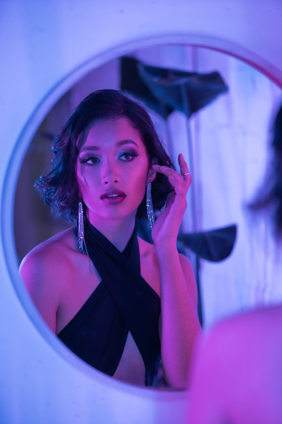 Stylish young brunette asian woman touching earring near mirror and neon light in night club - Foto, imagen