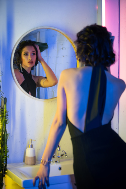 Модная юная брюнетка азиатка, прикасающаяся к волосам возле зеркала и тонущая в неоновом свете в ночном клубе - Фото, изображение