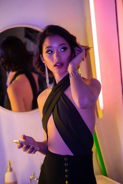 Trendy young asian woman holding smartphone and touching hair in neon light in night club - Foto, imagen