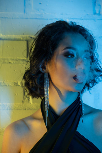 Portrait of fashionable young asian woman with makeup exhaling smoke in neon light in night club - Фото, изображение