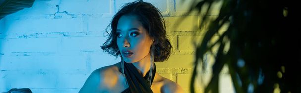 Stylish young asian woman looking away and standing near plants in neon light in night club, banner - Photo, Image