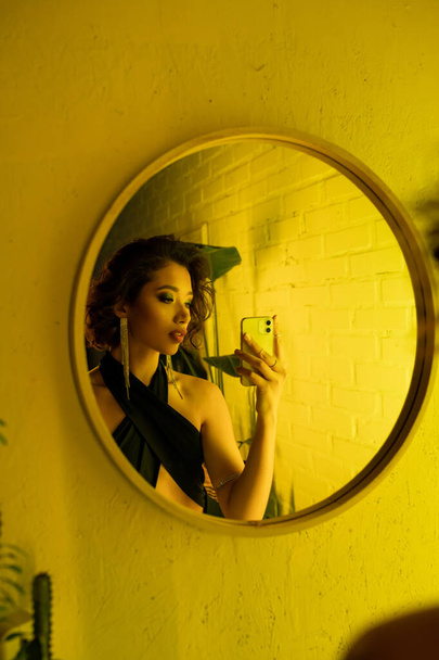 Stilvolle asiatische Frau im Kleid macht Selfie in der Nähe von Spiegel und gelbe Beleuchtung in Nachtclub - Foto, Bild