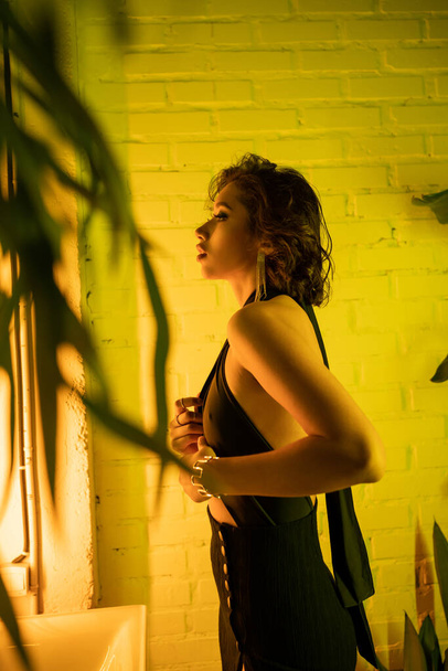 Widok z boku elegancka azjatycka kobieta dotykając sukienka stojąc w żółtym świetle neon w nocnym klubie - Zdjęcie, obraz