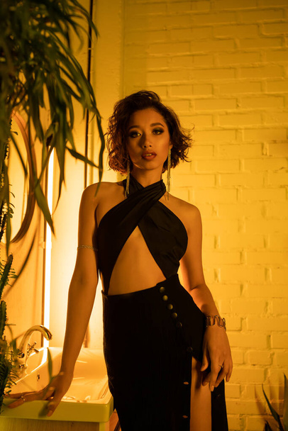 Sexy aziatische vrouw in jurk staan in de buurt gootsteen en plant in nachtclub met neon verlichting - Foto, afbeelding