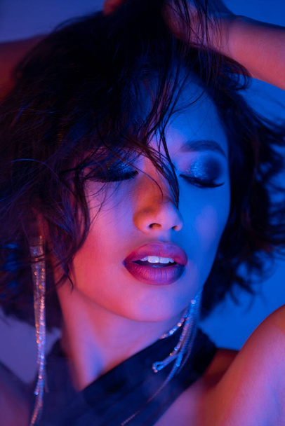 Elegante mulher asiática com penteado e maquiagem posando com olhos fechados em néon luz na boate - Foto, Imagem