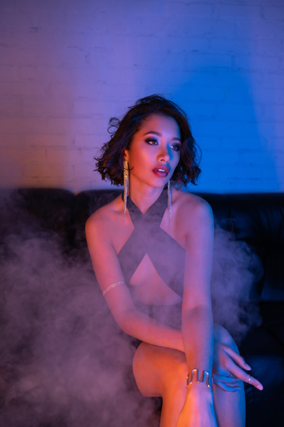 Elegant young asian woman in evening wear sitting in smoke and neon light on couch in night club - Foto, Bild