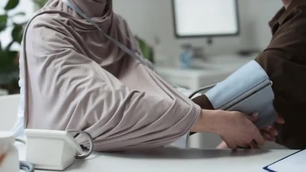 Close-up van jonge moslim vrouwelijke arts of huisarts in hijab controleren van de bloeddruk van de patiënt, terwijl beide zitten door de werkplek van clinicus in het medisch kantoor - Video