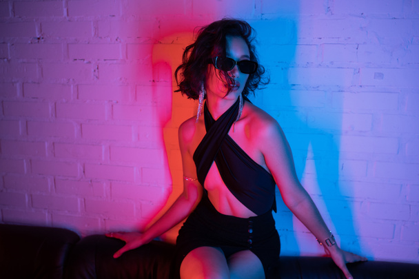Sexy asian woman in sunglasses posing in pink and blue neon light on couch in night club - Photo, Image
