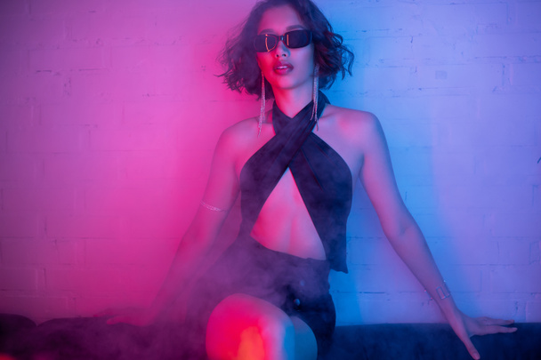 Fashionable asian woman in sexy dress and sunglasses posing in smoke and neon light in night club - Photo, Image