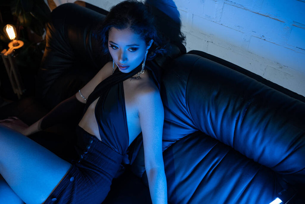 alto ângulo vista de elegante jovem asiático mulher sentado no sofá no azul néon luz no noite clube - Foto, Imagem