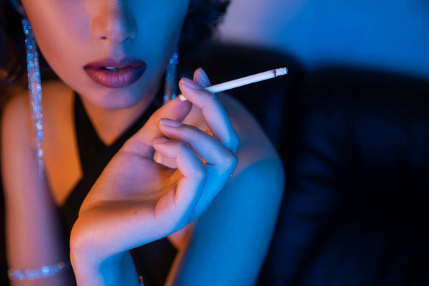 ナイトクラブでネオンライトに座っている間にタバコを保持するドレスでスタイリッシュな女性のクロップビュー - 写真・画像