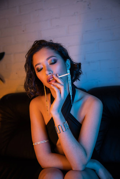 Élégant et élégant asiatique femme fumer cigarette tout en passant du temps dans la boîte de nuit avec néon lumière - Photo, image